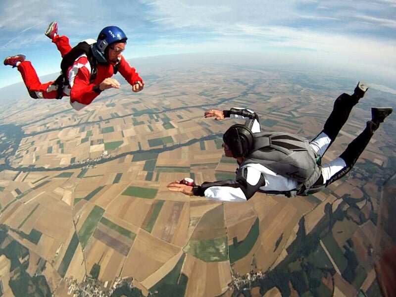 Deux parachutistes en chute libre, combinaison rouge et noire, survolant un paysage de champs agricoles.