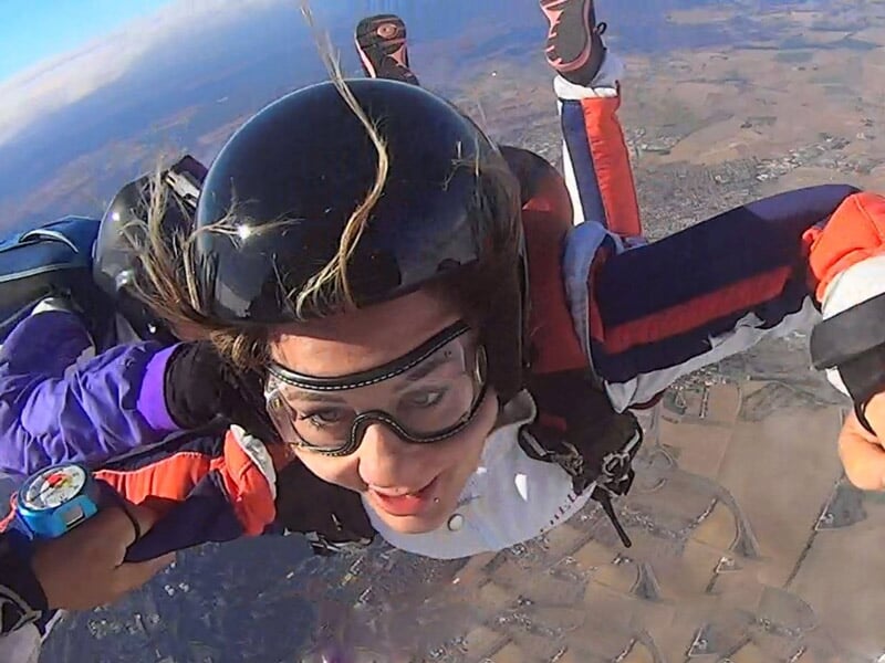 Femme en chute libre avec casque et lunettes, accompagnée d'un instructeur, paysage rural en dessous.