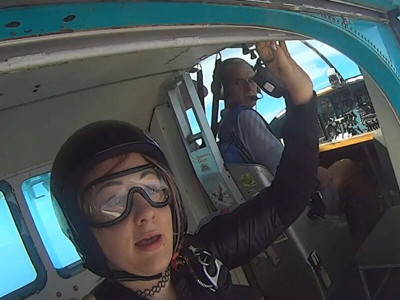 Personne équipée d'un casque et de lunettes de protection, prête à sauter d'un avion. Le pilote est visible dans le cockpit en arrière-plan, tenant une commande, avec un décor intérieur d'avion de sau