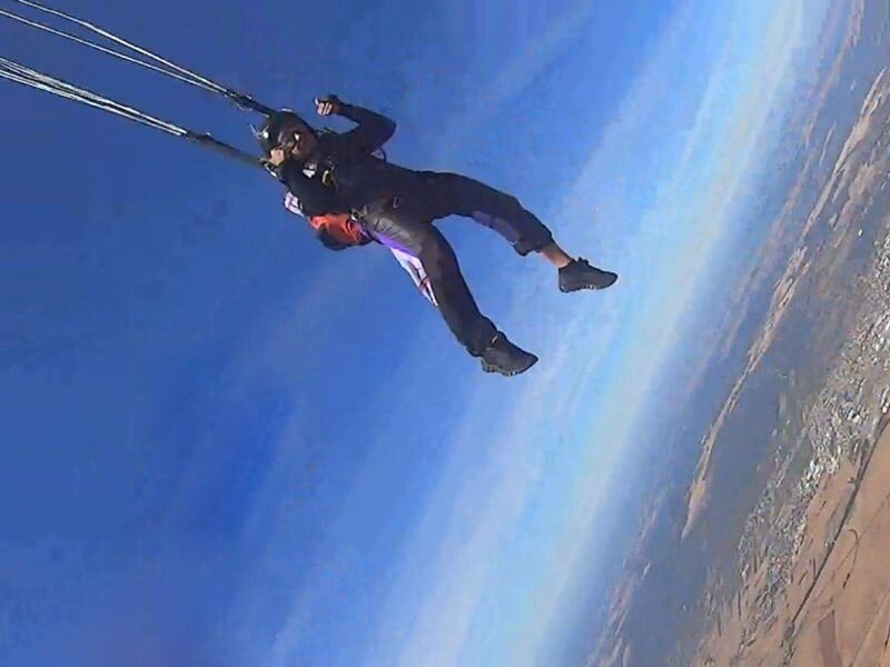 Personne en plein saut en parachute sous un ciel bleu dégagé, les suspentes du parachute visibles au-dessus. Vue sur un paysage en contrebas avec des terres agricoles et des zones urbaines, montrant l