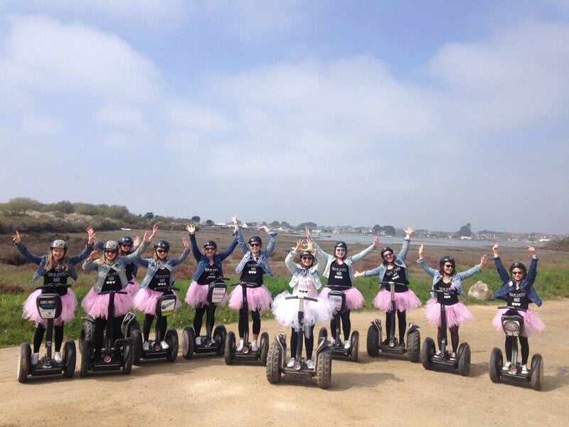Groupe de personnes en segway avec jupes roses et blanches, posant joyeusement en plein air près d'un plan d'eau.