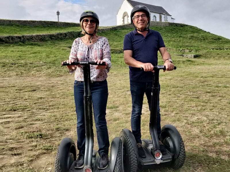 Un couple souriant en segway sur une pelouse verte avec une petite chapelle blanche en arrière-plan.