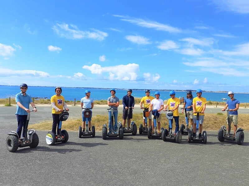 Groupe d'hommes en segway sur une route côtière, avec la mer et un ciel bleu en arrière-plan.