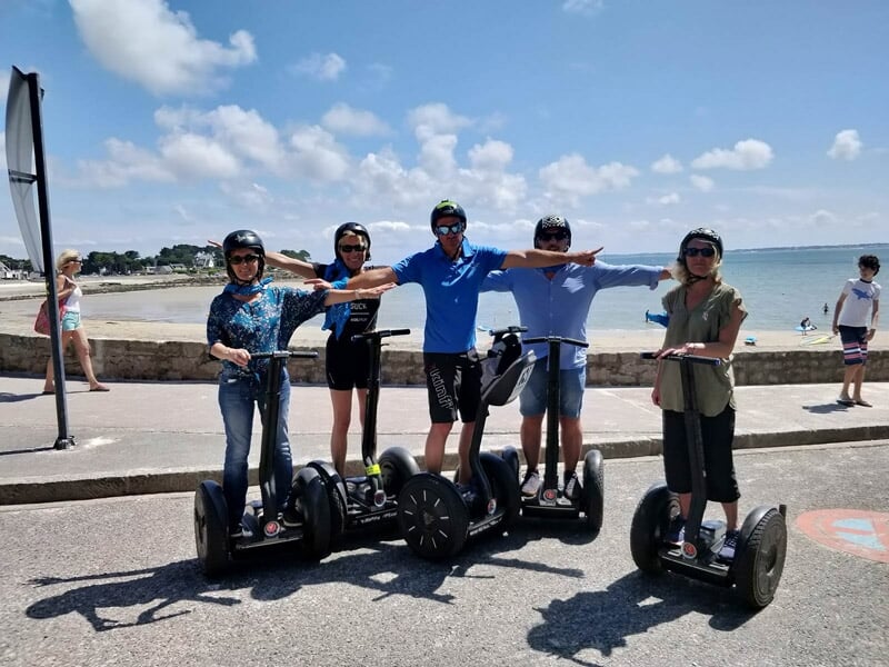 Cinq personnes en segway posant sur un quai au bord de la mer, bras tendus en avant, sous un ciel clair.