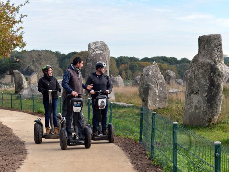 Trois hommes en segway discutant près d'un site de menhirs entouré d'une clôture verte.
