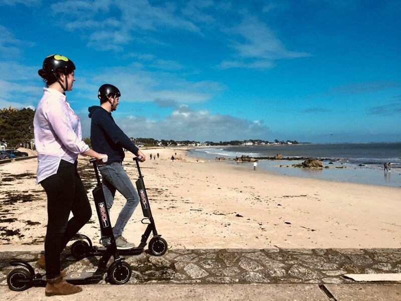 Deux personnes en trottinette électrique sur un chemin de bord de mer, regardant vers l'océan.