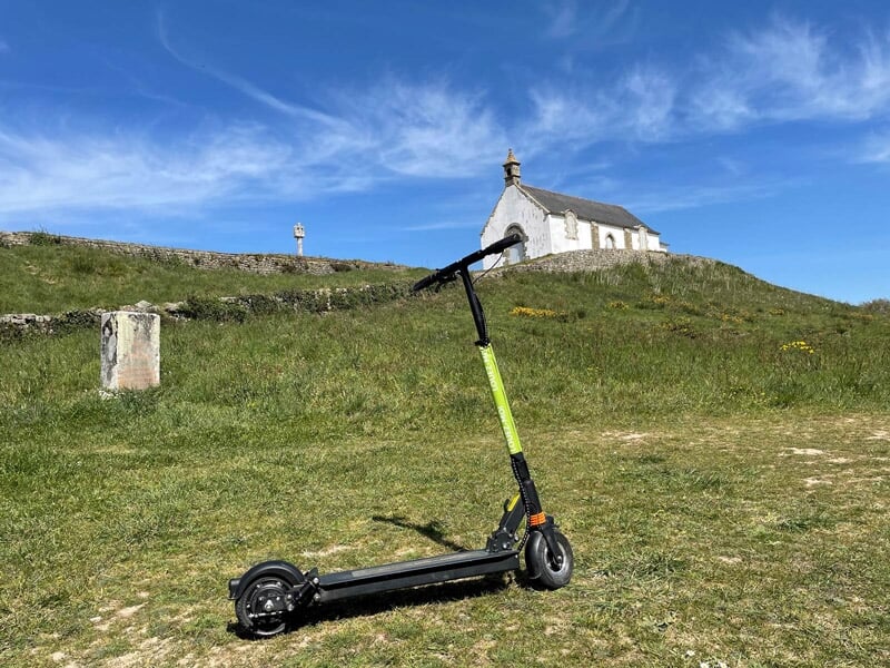 Trottinette électrique noire et verte posée sur une colline herbeuse avec une chapelle en arrière-plan.