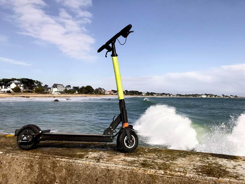 Trottinette électrique noire et verte posée sur un muret en bord de mer avec des vagues et un ciel bleu.