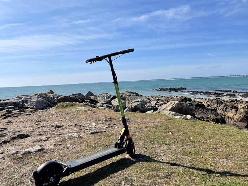 Trottinette électrique noire et verte posée sur un rocher au bord de la mer turquoise sous un ciel clair.