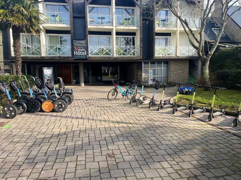 Alignement de segways et trottinettes électriques devant un hôtel avec des arbres et un ciel bleu.