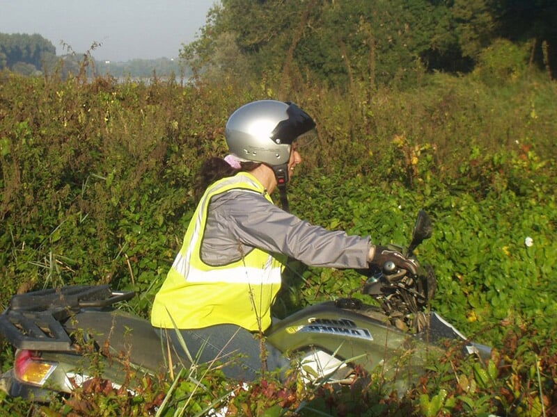 Personne en gilet jaune et casque argenté conduisant un quad dans un champ avec des buissons verts autour.