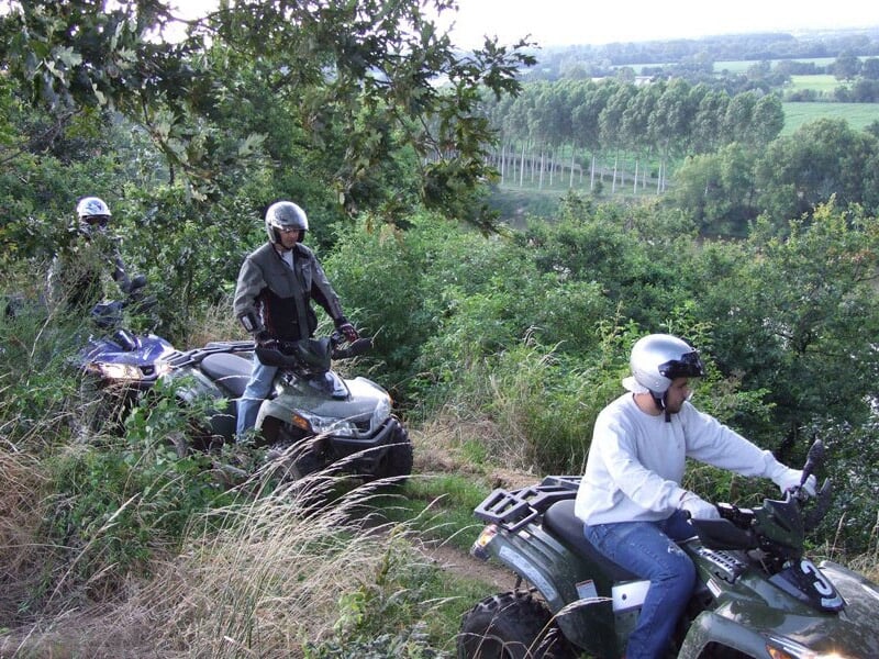 Trois personnes en casque sur des quads traversant un chemin herbeux bordé d'arbres et de buissons verts.