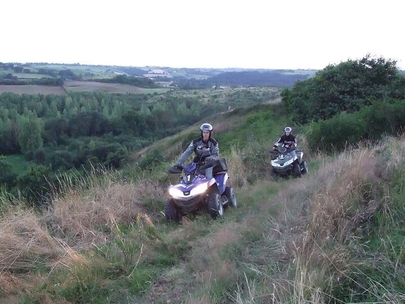 Deux conducteurs de quad en casque roulant sur un sentier herbeux avec une vue panoramique sur la campagne.