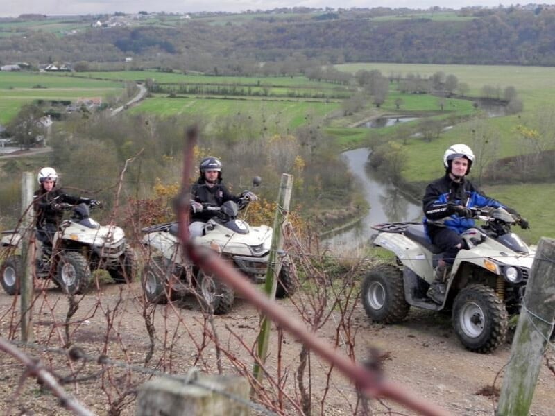 Quatre personnes en quad sur un chemin de terre avec une vue sur des champs et une rivière en arrière-plan.