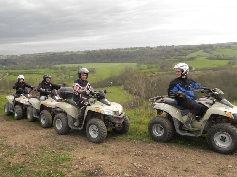 Quatre conducteurs de quad en ligne sur un chemin de terre dans un paysage vallonné et verdoyant.