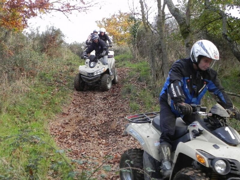 Trois conducteurs de quad en casque roulant sur un sentier étroit couvert de feuilles mortes en forêt.