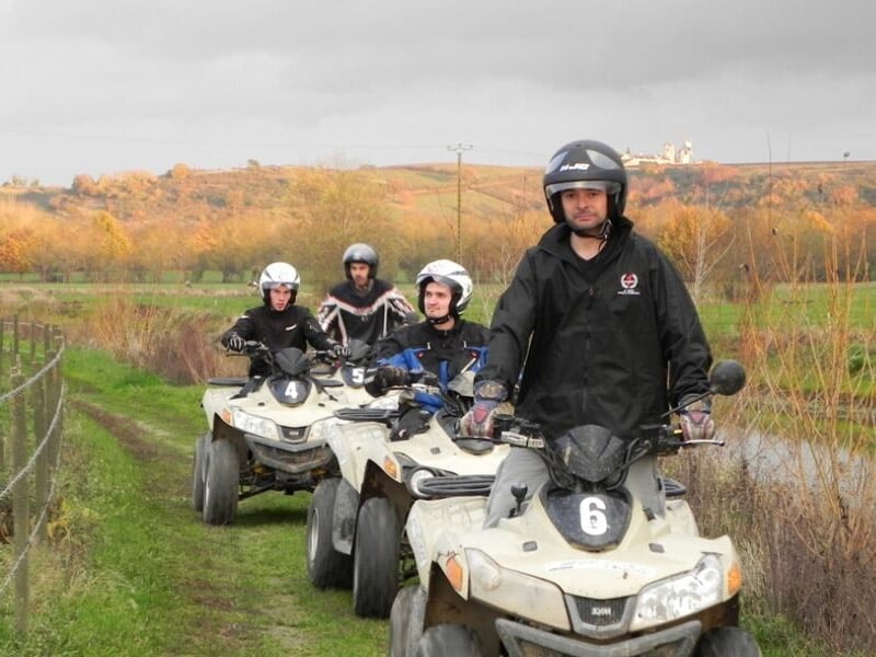 Quatre personnes en quad sur un chemin de campagne avec des champs verts et un ciel nuageux en arrière-plan.