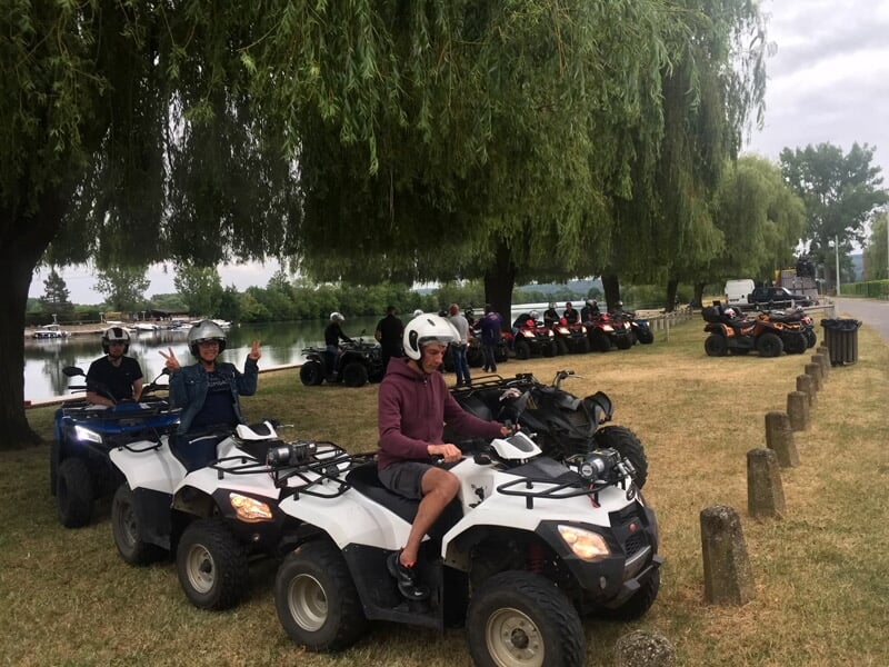 Personnes en casque sur des quads blancs alignés près d'un plan d'eau et sous un grand saule pleureur.