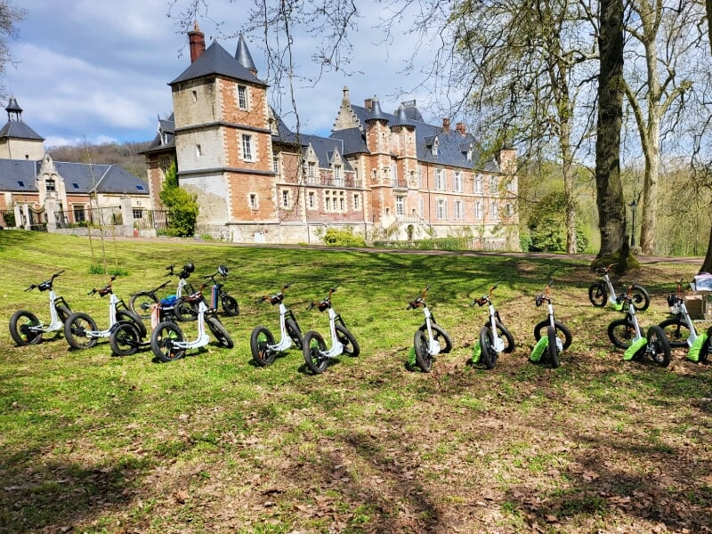 Alignement de trottinettes électriques noires sur une pelouse devant un château en briques sous un ciel bleu.