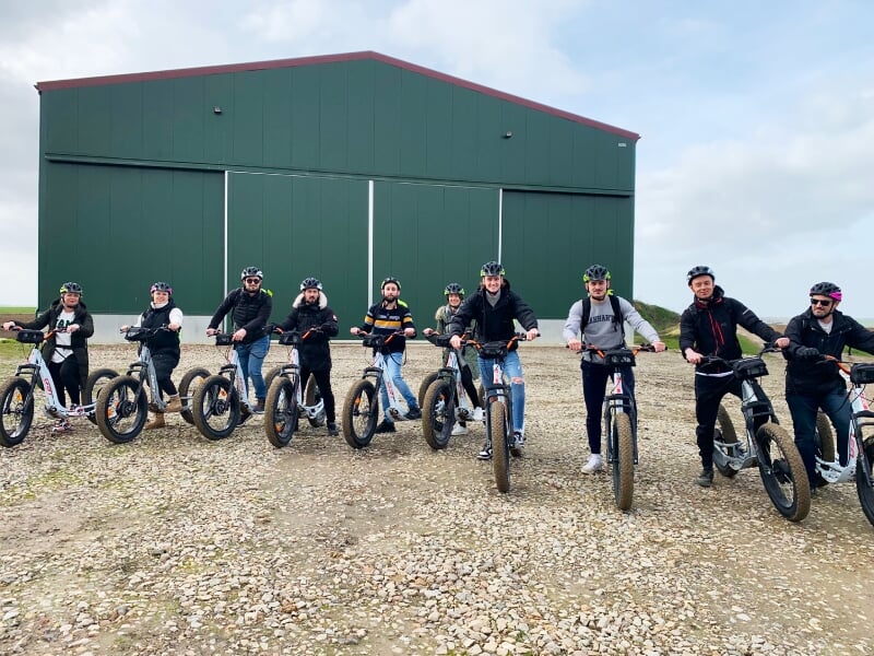 Groupe de personnes sur des trottinettes électriques noires posant devant un grand bâtiment vert industriel.
