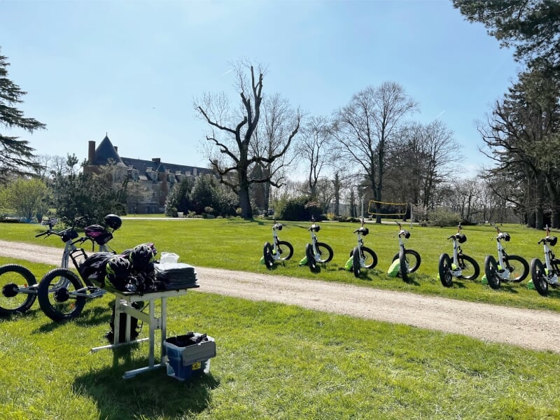 Trottinettes électriques noires alignées sur une allée herbeuse avec un château en arrière-plan au printemps.