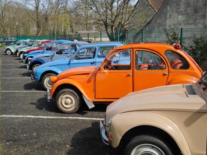 Plusieurs Citroën 2CV de différentes couleurs alignées dans un parking, avec une 2CV orange vif au premier plan, suivie de modèles bleus et beiges. Le fond montre des arbres sans feuilles et une clôtu