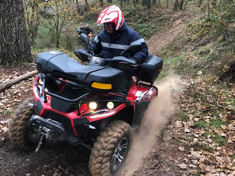 Conducteur en casque rouge sur un quad rouge traversant un sentier forestier boueux avec des feuilles au sol.
