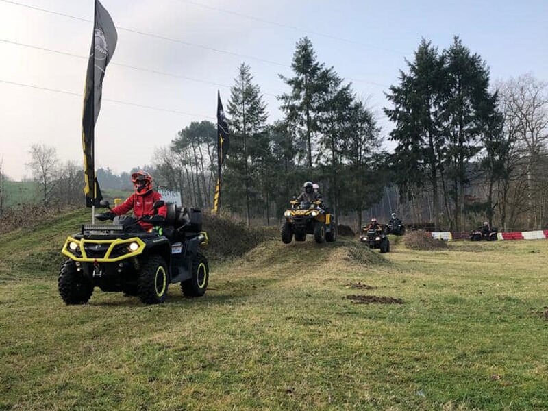 Plusieurs quads roulant sur une piste herbeuse avec des arbres et des drapeaux noirs en arrière-plan.