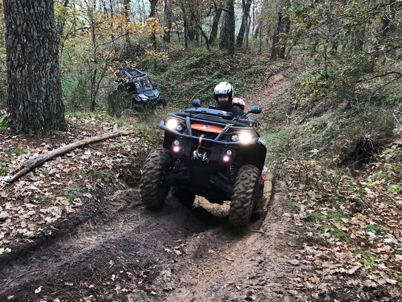 Quad noir avec conducteur en casque blanc roulant sur un chemin forestier boueux entouré d'arbres.