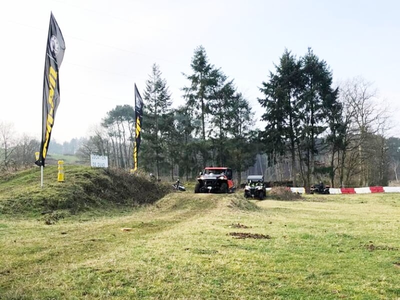 Quads roulant sur une piste herbeuse avec des bosses, des arbres et des drapeaux noirs visibles.