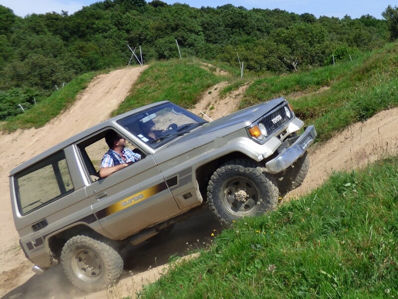 Un 4x4 gris monte une pente terreuse en terrain accidenté avec un conducteur concentré au volant.