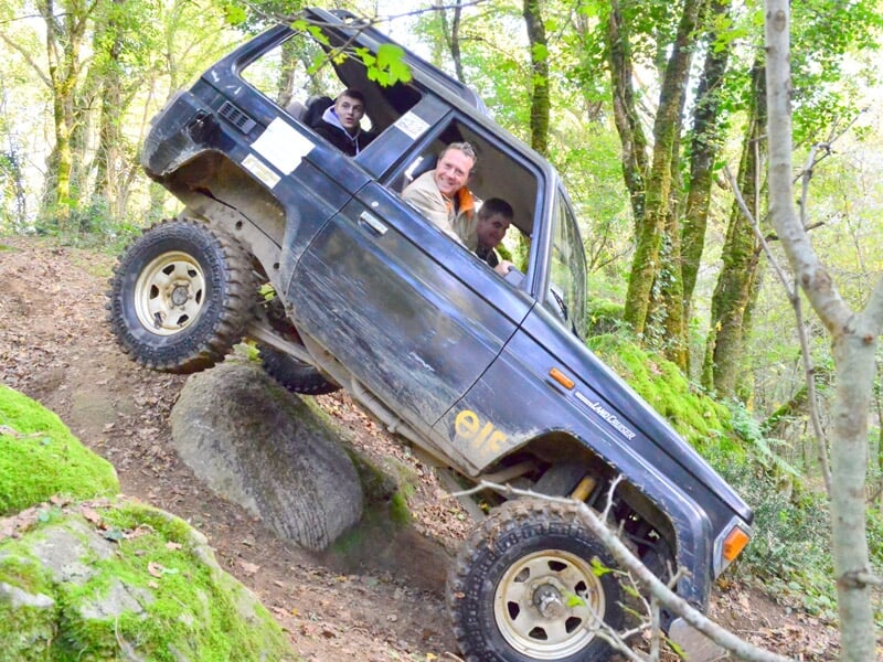 Un 4x4 noir avec plusieurs passagers franchit un obstacle rocheux en forêt dense.