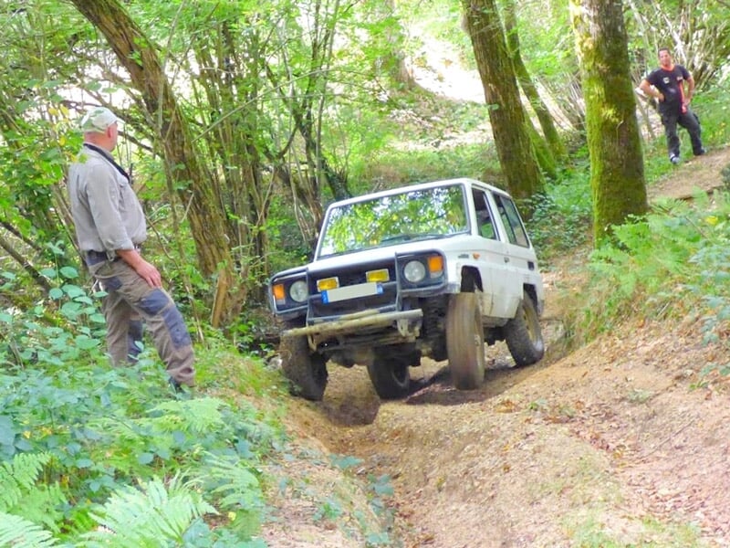 Un 4x4 blanc évolue sur un sentier forestier étroit avec deux personnes observant à proximité.
