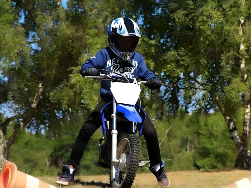 Un jeune enfant en tenue de motocross bleue manœuvre une petite moto sur un parcours d'entraînement avec des cônes sur l'herbe.