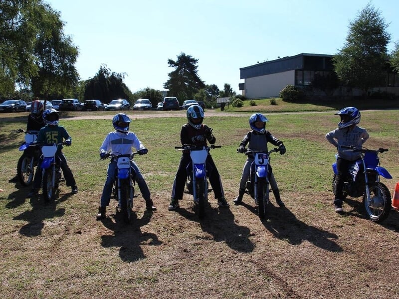 Cinq jeunes motocyclistes en tenue complète de motocross alignés et prêts à commencer une session d'entraînement sur un terrain herbeux.