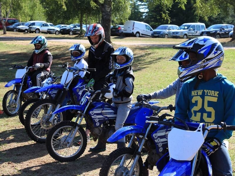 n groupe d'enfants portant des équipements de sécurité pour motocross, y compris des casques et des gilets, assis sur des motos dans un champ avec des voitures en arrière-plan.
