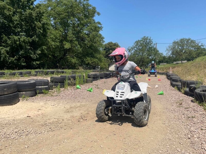 Enfant en casque rose conduisant un quad blanc sur une piste de terre entourée de pneus et de verdure.