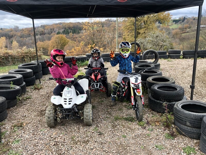 Trois enfants en tenue de protection posant avec un quad et deux motos sur un terrain de course en gravier.