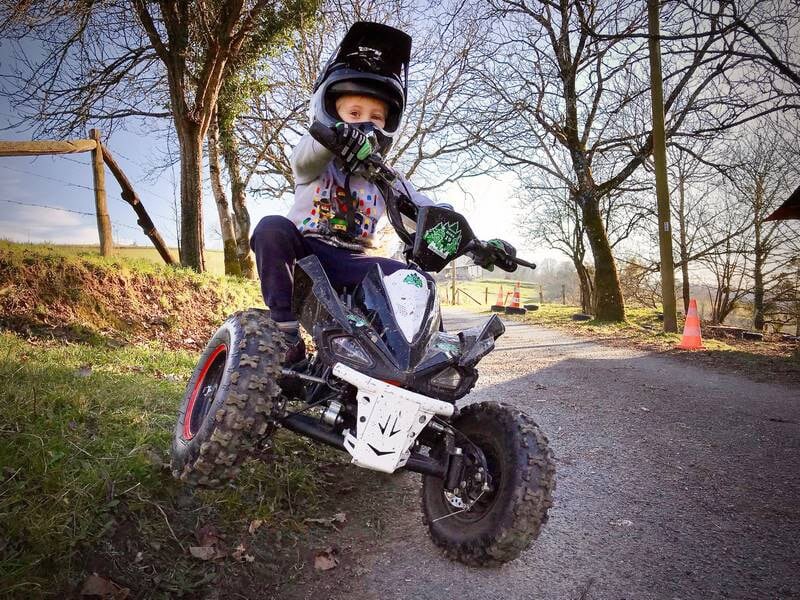 Jeune enfant en casque noir conduisant un petit quad électrique sur un chemin en terre entouré d'arbres.