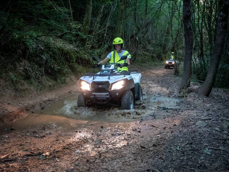 Personne en gilet jaune conduisant un quad tout-terrain dans une zone boueuse en forêt avec phares allumés.