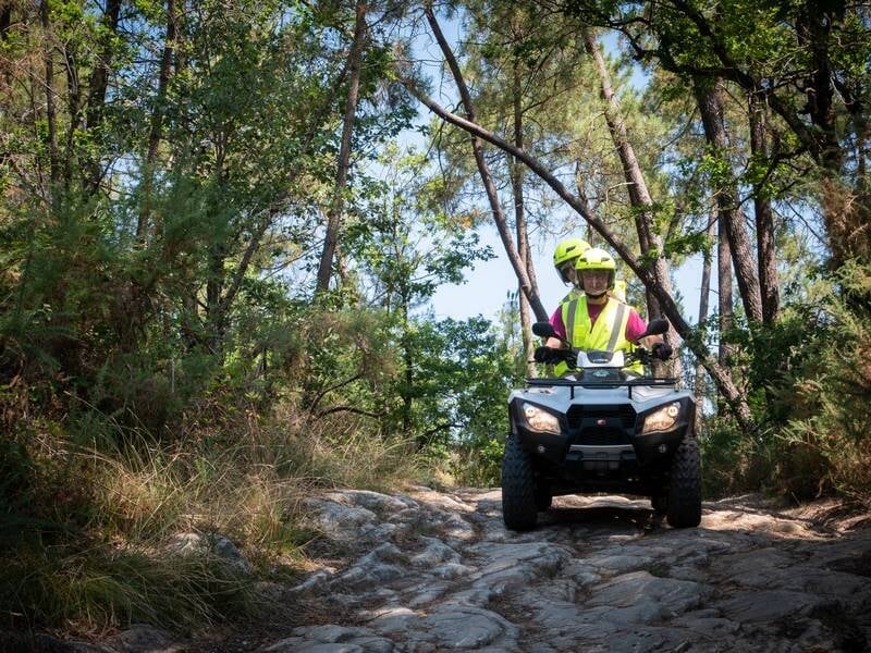Personne souriante en casque vert conduisant un quad sur un chemin rocheux en forêt sous un ciel clair.