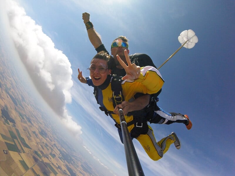 Deux personnes en saut en parachute au-dessus d'un paysage champêtre avec ciel bleu et nuages blancs.
