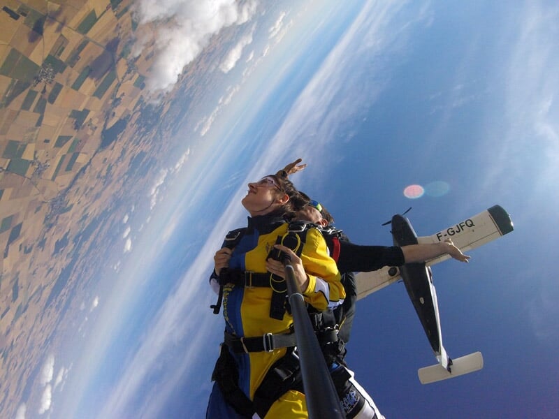 Parachutistes en chute libre avec avion en arrière-plan et ciel dégagé au-dessus des champs.