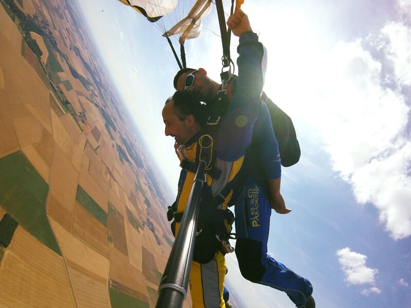 Parachutistes en tandem en plein vol avec vue sur champs et ciel partiellement nuageux.