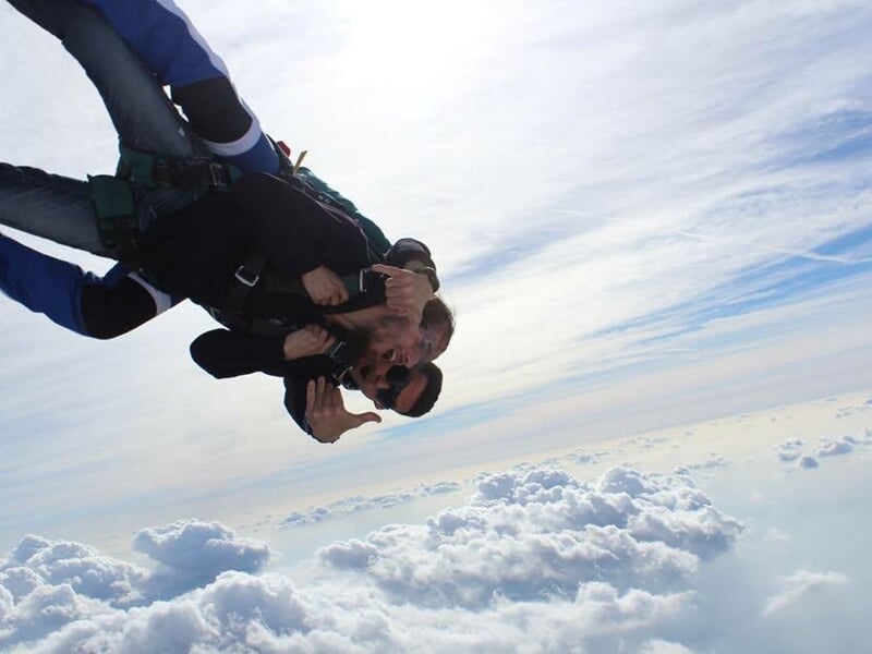 Saut en parachute en tandem au-dessus des nuages, deux personnes avec pouce levé et ciel clair.