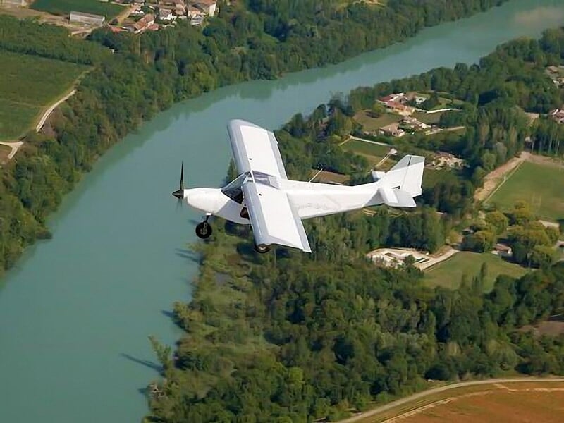 Avion blanc survolant une rivière verte sinueuse entourée de champs et villages verdoyants vus d’en haut.