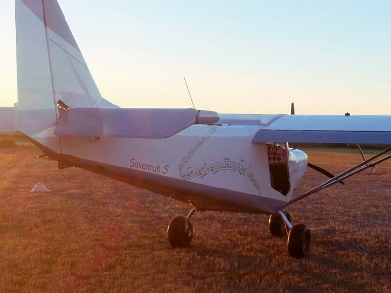 Avion léger blanc marqué « Savannah S » stationné sur herbe sèche, décoré de notes de musique au fuselage.