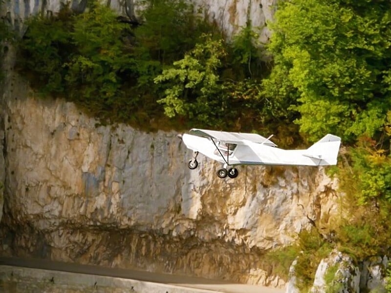 Petit avion blanc en vol rasant une falaise rocheuse bordée d’arbres verts sous une lumière chaude.