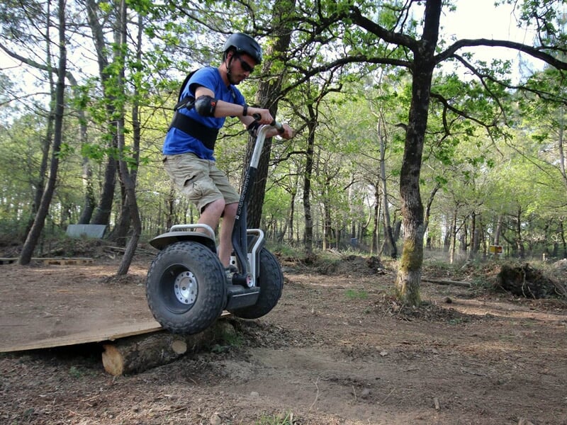 Homme en casque bleu sur un gyropode tout-terrain franchissant un obstacle en bois dans une forêt verdoyante.
