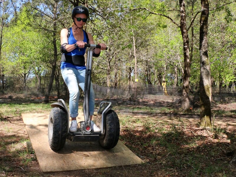Femme en lunettes de soleil et casque noir sur un gyropode roulant sur une planche en bois dans un bois.